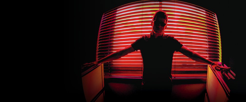 Noel Gallagher stands confidently in front of illuminated blinds
