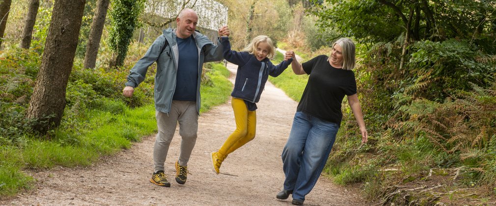 A family walking on a wide forest trail. The couple are swinging their daughter in the air.