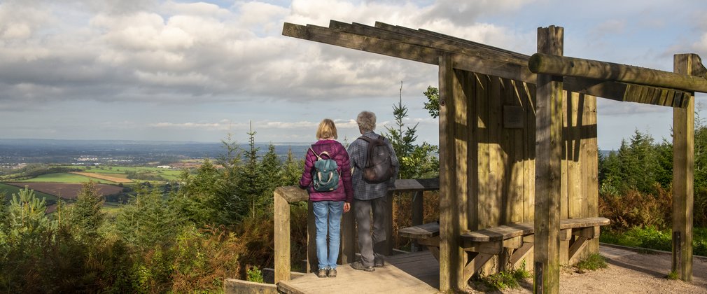 A couple stand under a wooden shelter looking out from a viewpoint across the countryside.