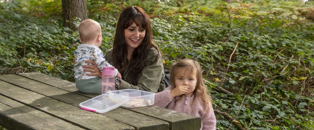A woman with a baby and small child enjoying lunch at a picnic table in a forest