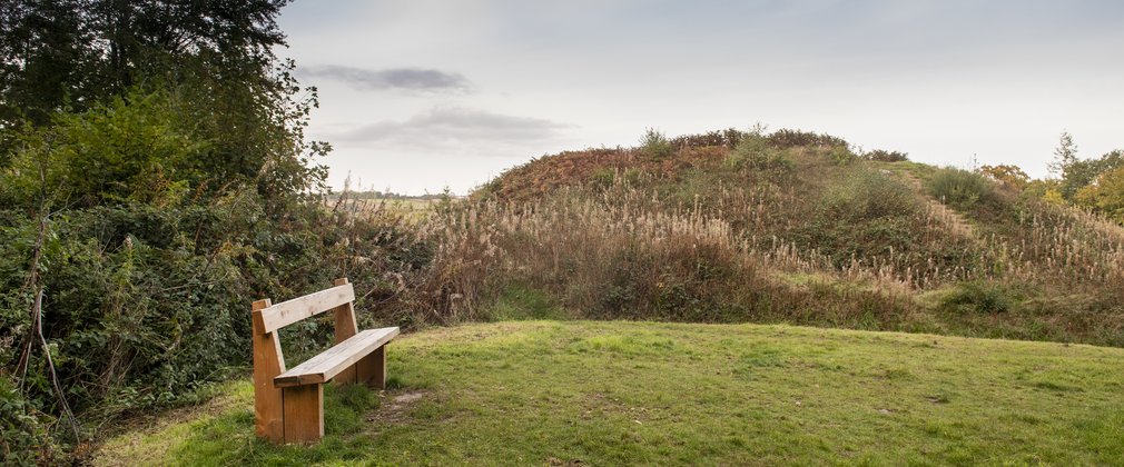A motte and bailey monument viewed with a grassy area and wooden bench in the foreground