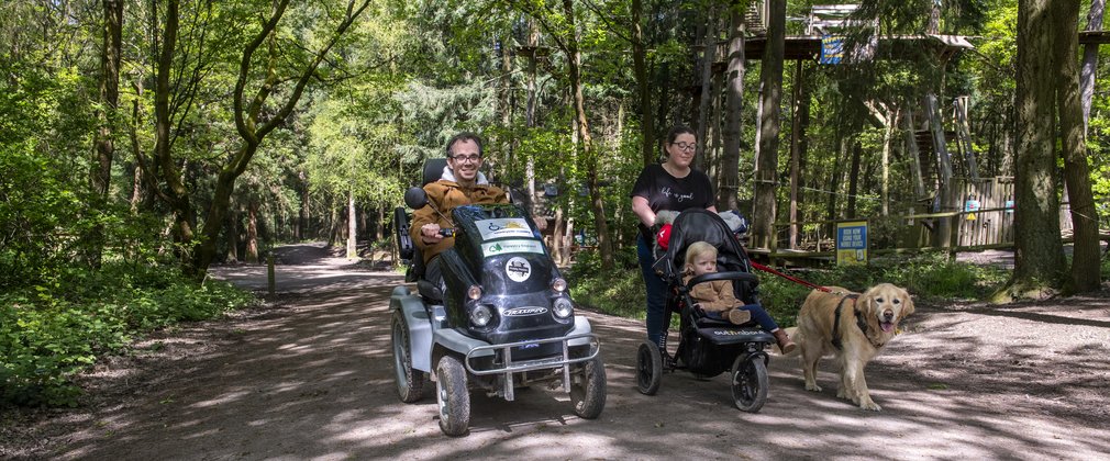 Man using mobility scooter, alongside a woman pushing a pram and walking a dog