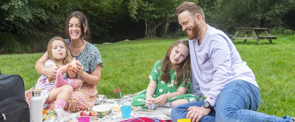 Two parents and two children sit on a tartan blanket having a picnic in a grassed area