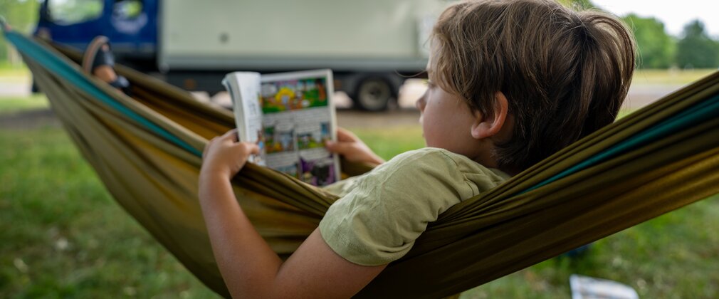 Boy reading a graphic novel whilst lying in a hammock, with a motorhome in the background