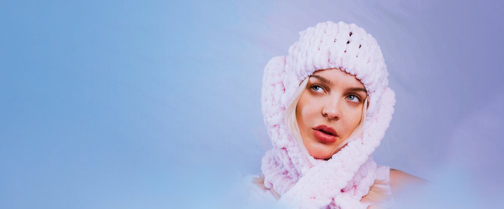 Anne-Marie wearing a knitted pink hat staring up to the left against a blue background