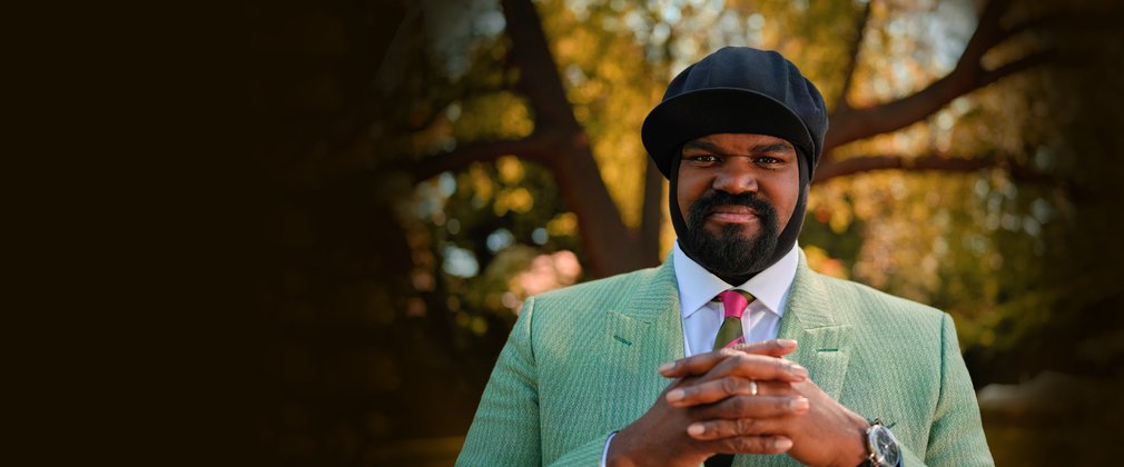 Gregory Porter in a green suit and hat stood in front of trees blurred in the background.