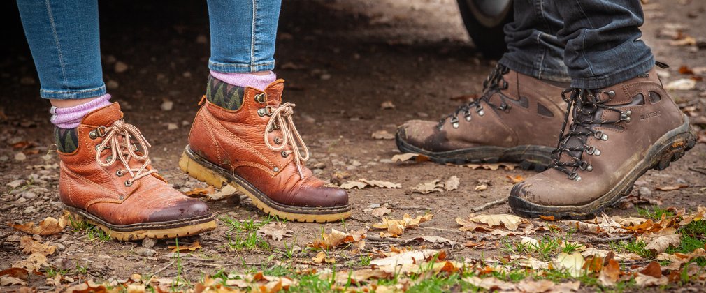 Close-up of two people's walking boots stood on forest ground.