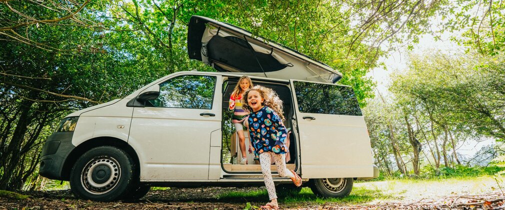 Two girls running out of a white campervan surrounded by trees