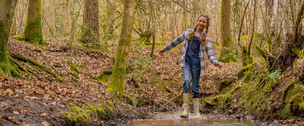 A girl wearing rubber boots splashing in a forest puddle.