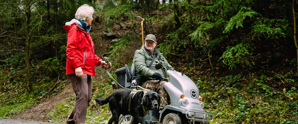 A man on a mobility scooter and a woman with a black dog are on a forest path