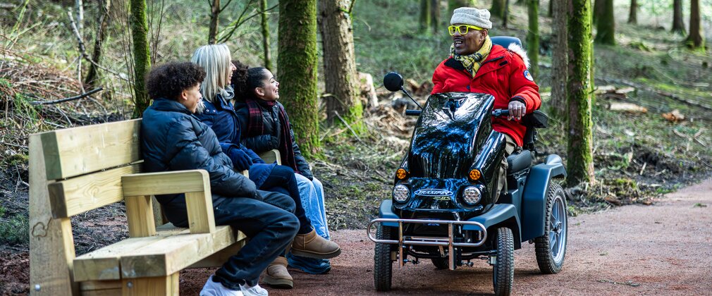 Family sitting on a bench in a forest, the dad is in a mobility scooter