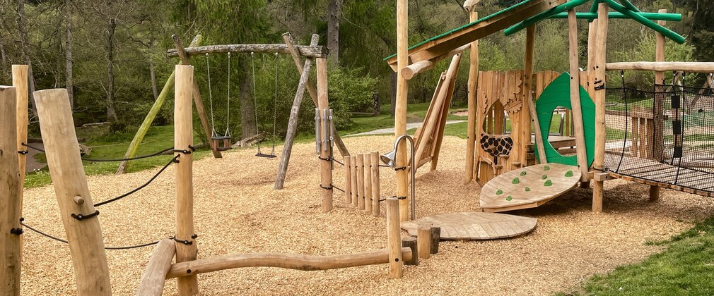A woodland play area with balance beams, a small climbing wall and swings.