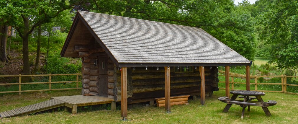 A log cabin in a grassed area surrounded by a wooden fence with trees behind