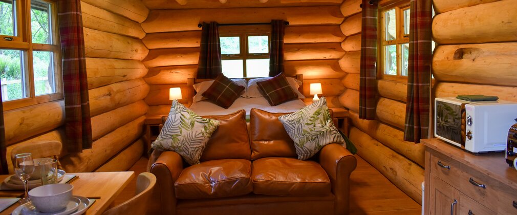 Interior of a rustic log cabin with bed, sofa and dining/kitchen area visible
