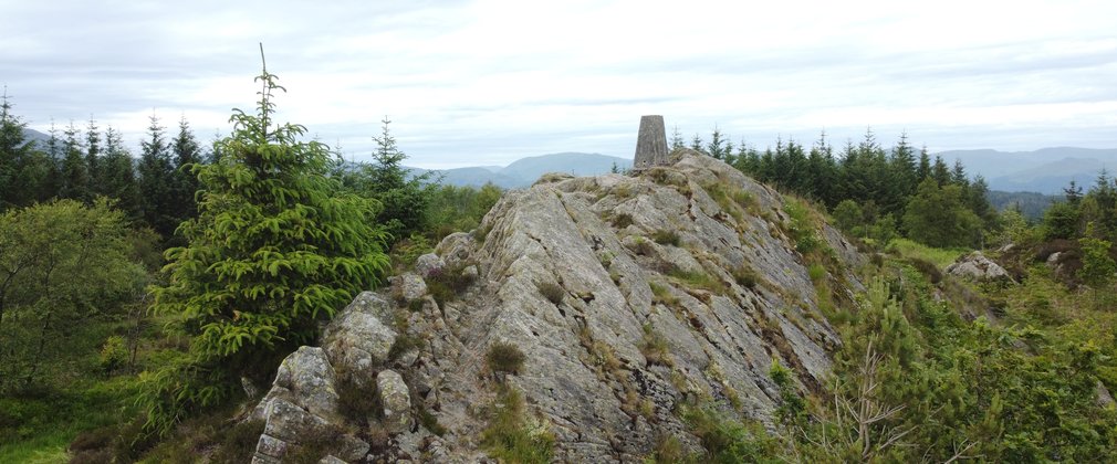 A trig point on top of a rocky outcrop above a forest