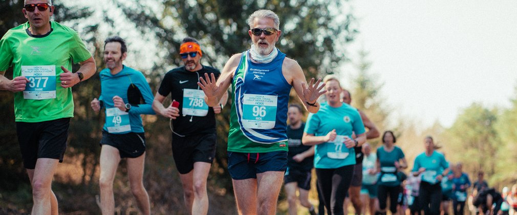 A line of runners running a long a hard path. A man in the foreground with sunglasses waves at the camera as he runs.