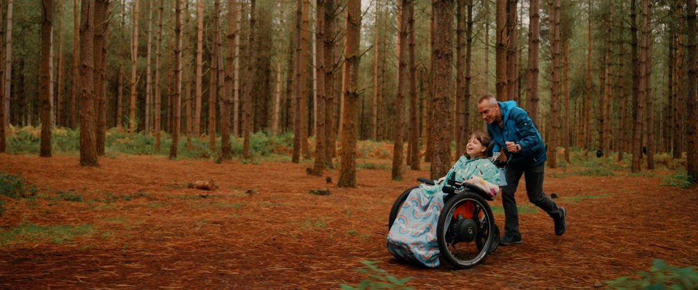 A young girl in a wheelchair smiling as a male carer pushes her through autumn woodland.