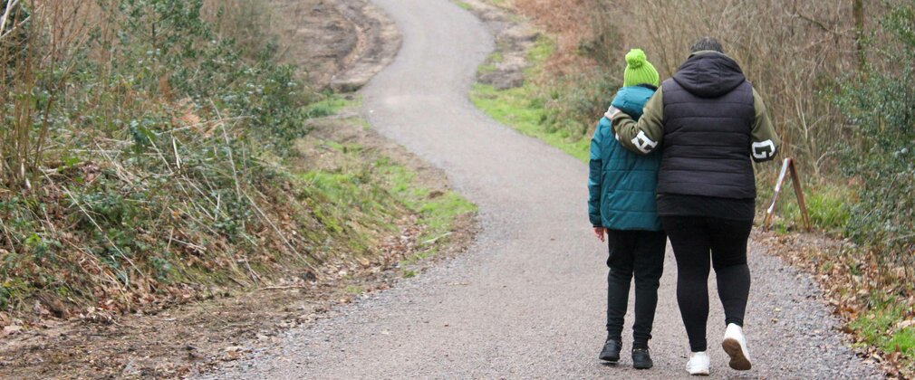 Two people walking along a winding gravel trail in the woods during winter