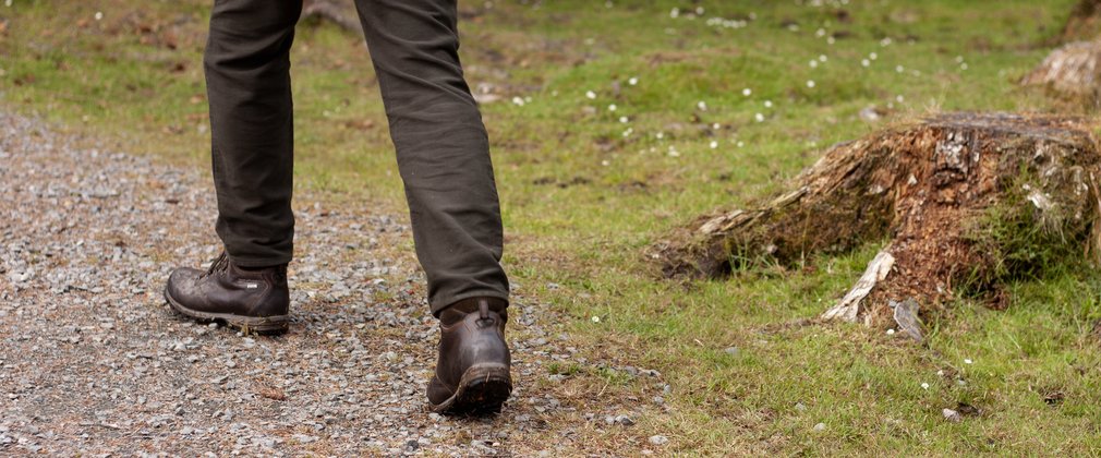 A close-up of a pair of legs walking on a gravel path through a forest.