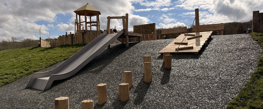 A playground of wood structures at the top of a small hill with a slide running down the hill