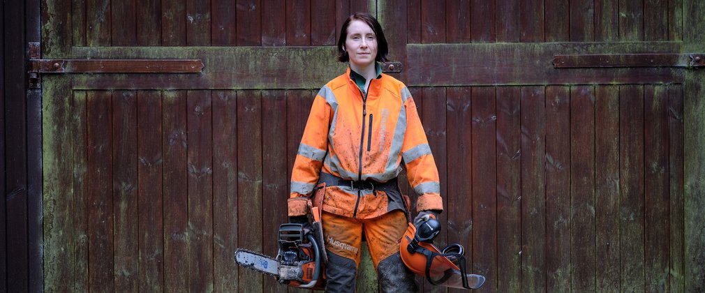 Woman stood in front of large wooden door with helmet and chainsaw in hand 