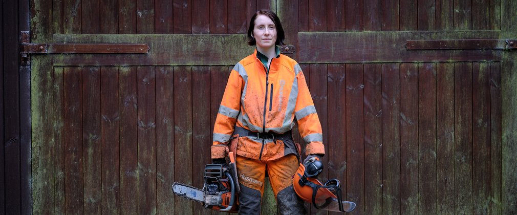 Woman stood in front of large wooden door with helmet and chainsaw in hand