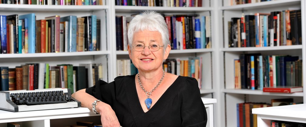Author Jacqueline Wilson smiles at the camera leaning on a book case covered in hundreds of books.
