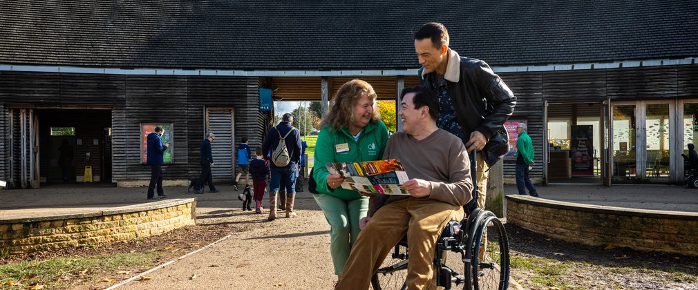 Two men, one standing and one in a wheelchair speaking to a lady in green in front of building whilst looking at a leaflet