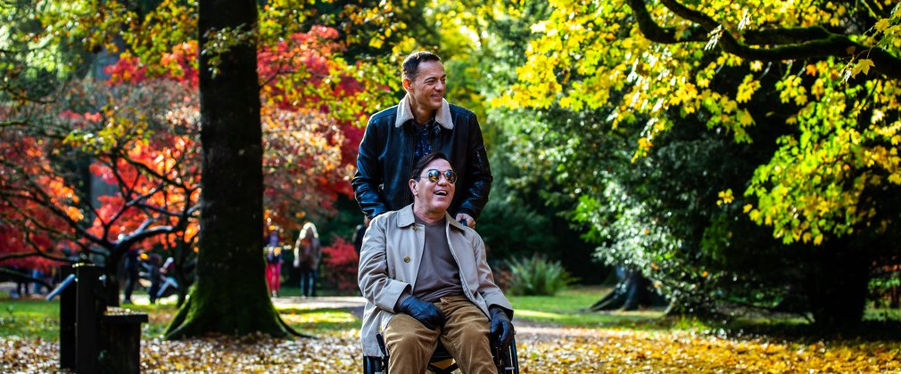 A man in a black jacket pushes a man in a wheel chair through a gorgeous autumnal scene