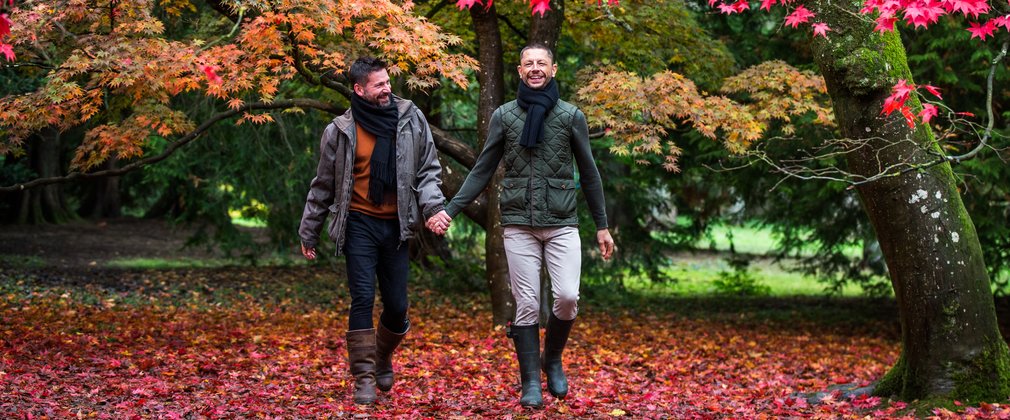 2 men walk hand in hand through a beautiful autumnal landscape of red and orange. Red leaves underfoot create an illusion of walking on a red carpet.