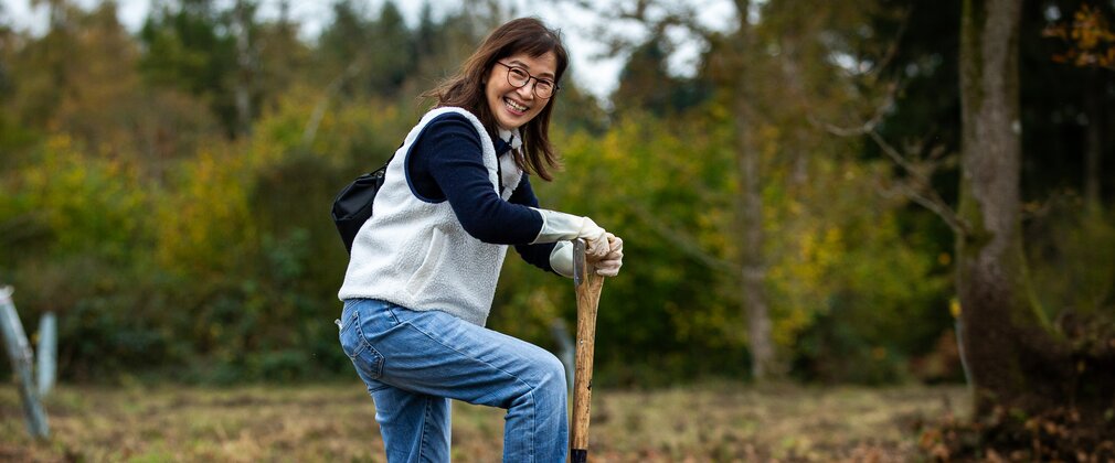 A woman smiles at the camera holding a shovel that she is pushing down on one leg into the soil.