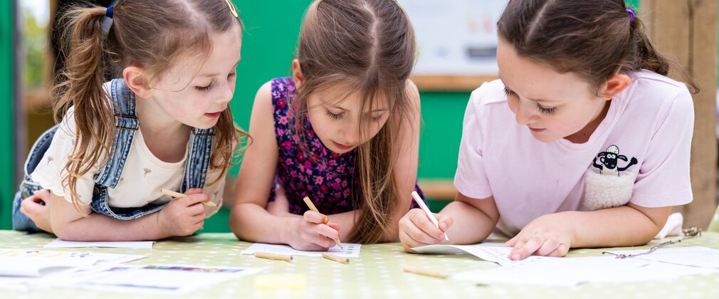 three young girls lean on a table to write and draw on paper together.