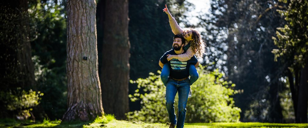 A young adult male carries a young adult female on his back as they playfully walk through a lush green opening in the trees. The young female adult points up to a feature in the distance.