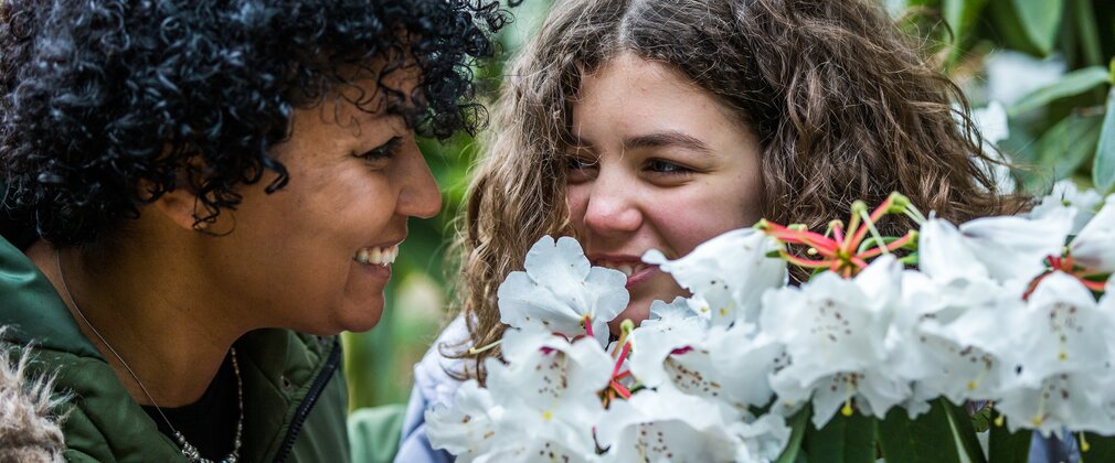 A mum and daughter smile at each other while smelling white rhododendron flowers