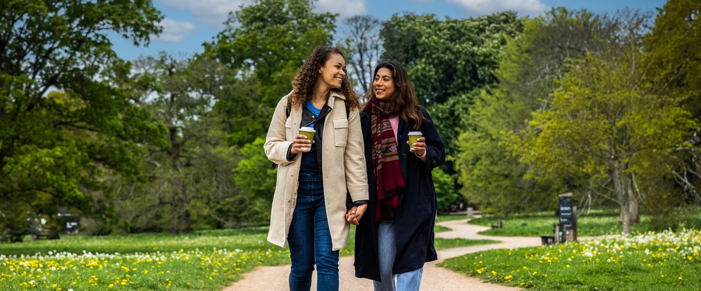 2 women walk hand in hand holding takeaway coffees cups. they smile at each other as they walk along a gravel path with lush green trees in the background.
