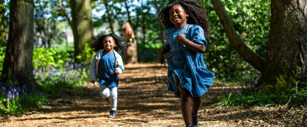 2 children run towards the camera smiling - they are running between trees with a sculpture of an owl in the background