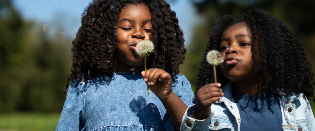 A young girl and boy dressed in denim hold dandelions up to their mouths to blow