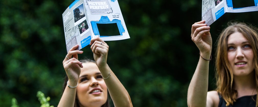 2 women hold picture perfect leaflets in their hands, it has a square cut out to look through. They hold them up to peer through