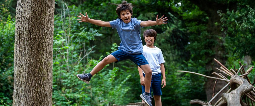A young boy in a blue t-shirt and dark blue shorts jumps high off a log with his arms and legs stretched out. His older brother stands behind him smiling.
