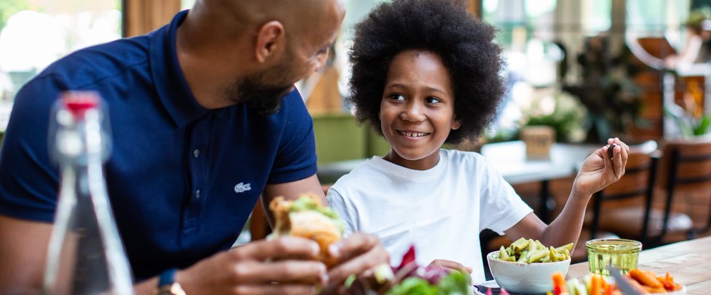 A father and son sit at a table with food in front of them. The father looks at his son who smiles back.