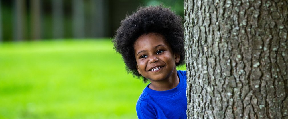 A young boy in a bright blue t-shirt pokes his head out from behind a tree trunk with a grin on his face.