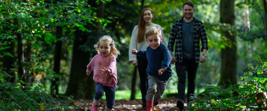 A young boy and girl run towards the camera over crunchy brown leaves with their parents smiling in the background.