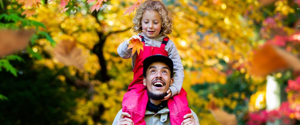 A boy sits on his dad shoulders holding a yellow maple leaf surrounded by autumn colour