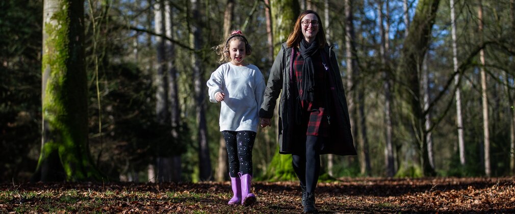 A woman and her niece stroll along a leaf strewn path with bare trees in the background smiling towards the camera.