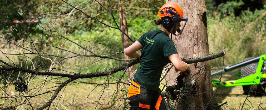 A woman in a green top and orage hard hat walks with a large branch towards a chipper