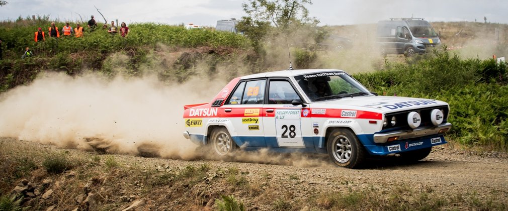 A white, blue and red rally car on a dirt track with dust billowing behind