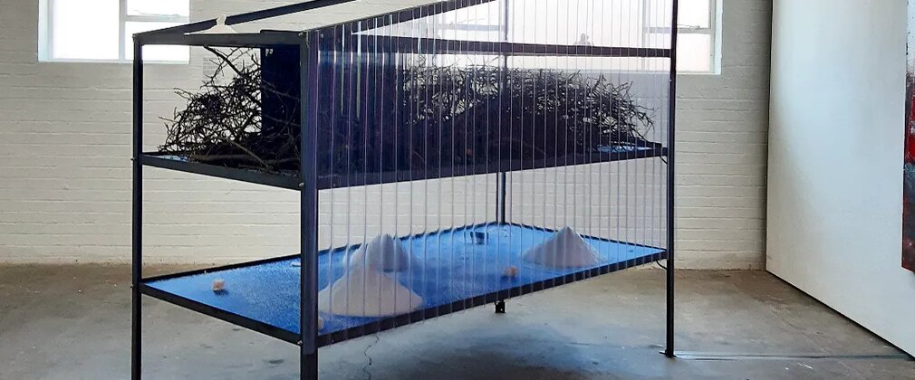 Contemporary installation featuring a black metal frame with suspended branches above a blue platform displayed in a bright industrial exhibition space.