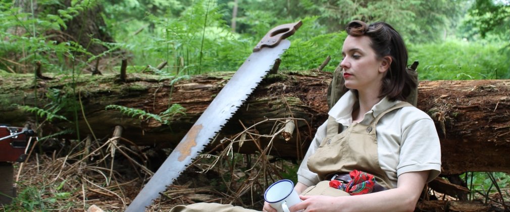An actress dressed as a Lumberjill. She is resting against a felled tree with a handsaw to her side. In her hand is an enamel white mug.