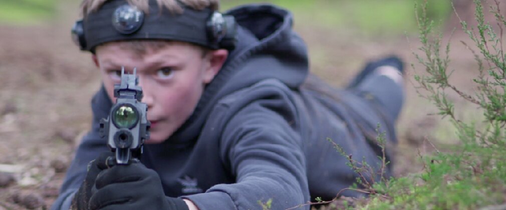 A child lying down in the forest pointing a toy laser gun 