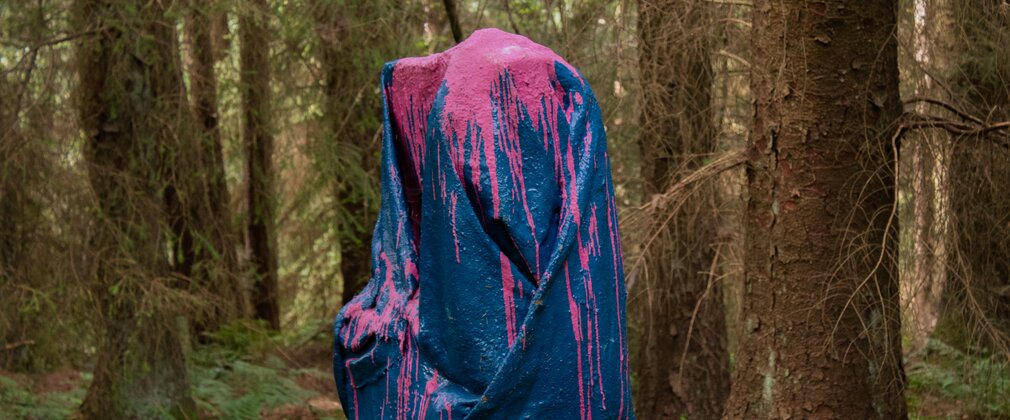 A mixed media sculpture that looks like a cloth stood up in the forest covered in blue and pink paint.